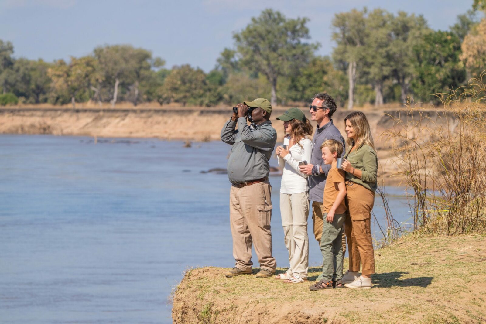 Image of a family with a young child and 1 guide on a walking safari in South Luangwa. The guide holds binoculars while the group overlooks the river. Courtesy of Time + Tide | TrueAfrica | Blog | Time + Tide
